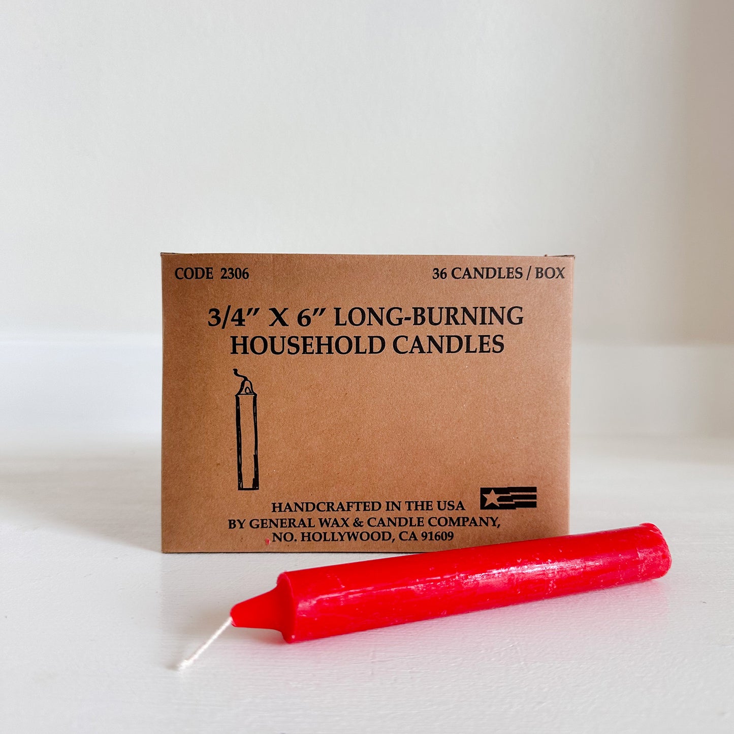 Red candle in front of a cardboard box labeled 'Long-Burning Household Candles' on a white background